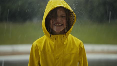 portrait of a happy teenage girl in a yellow jacket standing in the rain and catching raindrops with her hood in the park