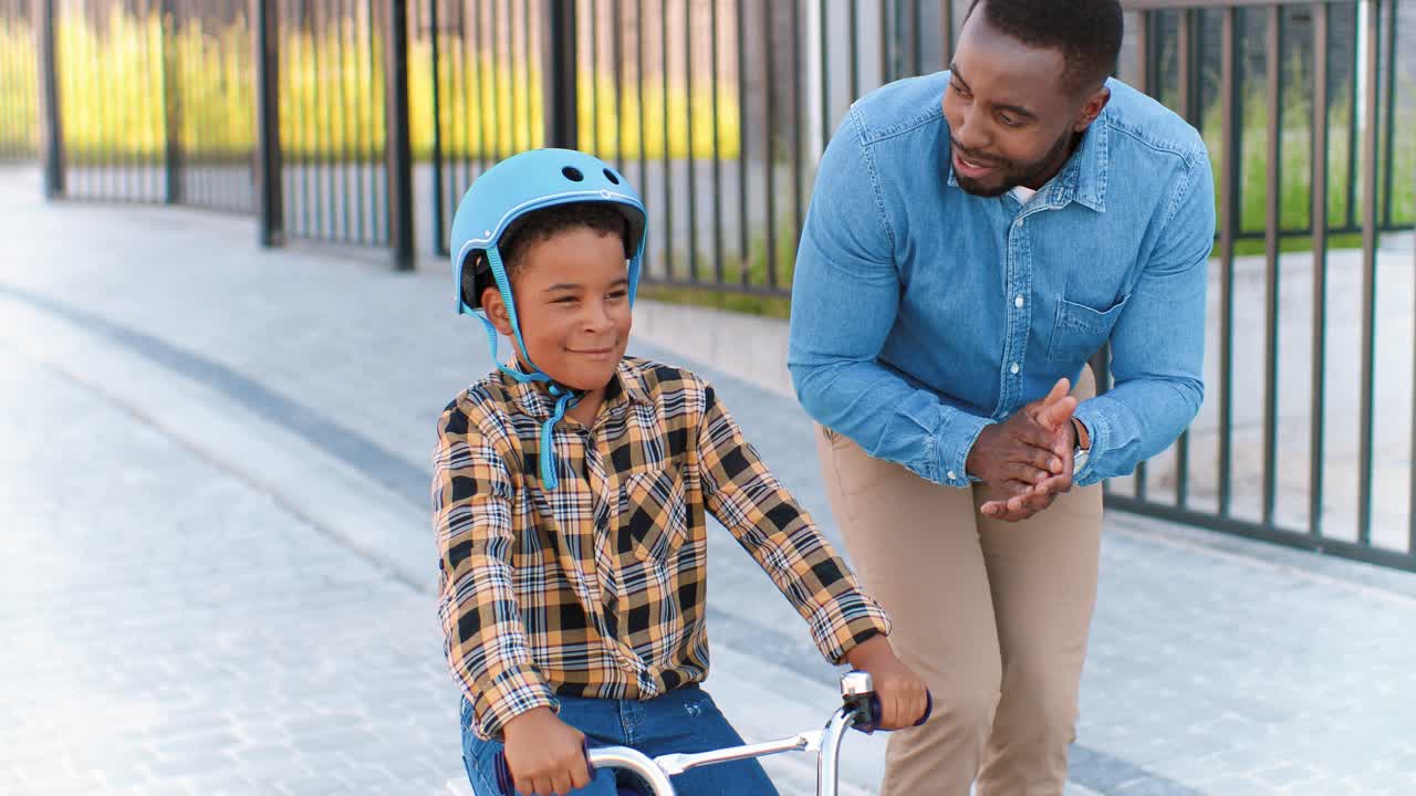 padre afroamericano enseñando a un niño pequeño con casco montando en bicicleta en la calle en el suburbio