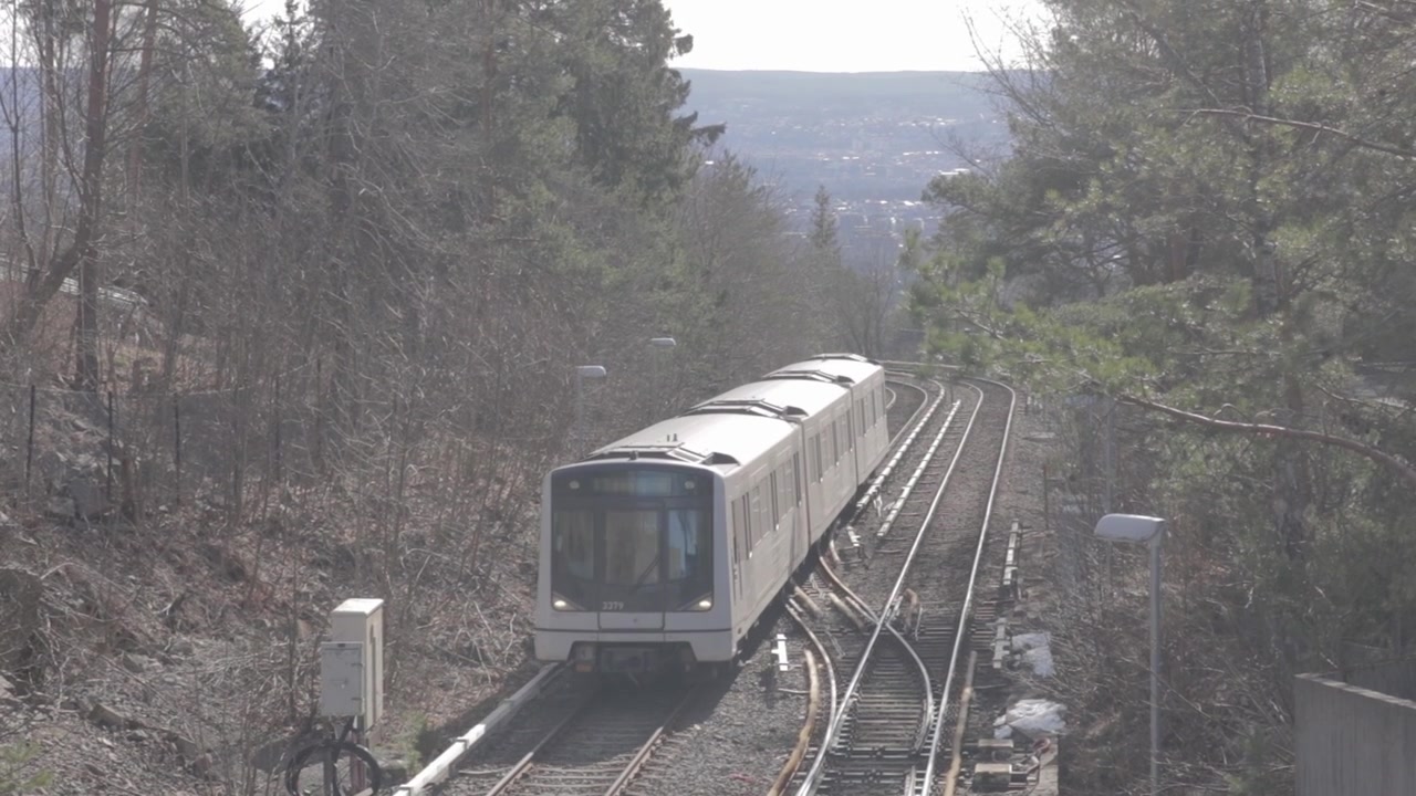 Pengambilan gambar dari ketinggian kereta Metro Oslo T-bane yang melaju di sepanjang rel luar ruangan, dibingkai oleh pohon pinus di lereng bukit menuju stasiun Frognerseteren, dengan pemandangan lembah yang jauh di Oslo, Norwegia
