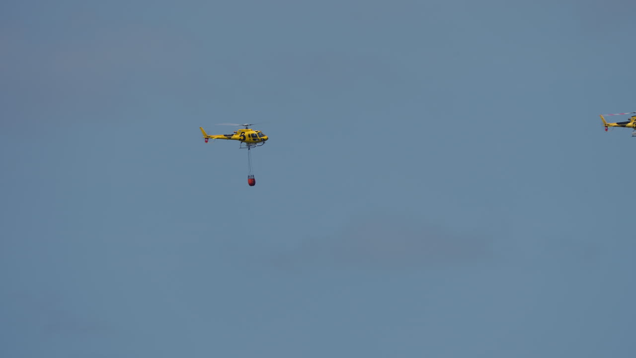 Yellow helicopter flying with suspended red bambi bucket used for firefighting purposes