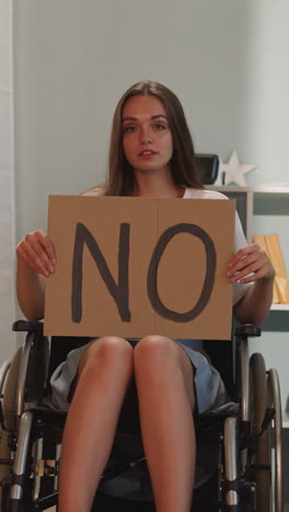 woman with disability shows poster with word no looking in camera. young female with paralysis protests against social inequality sitting in wheelchair at home