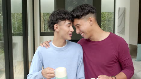 smiling multiracial gay couple at home, one holding coffee mug, enjoying time