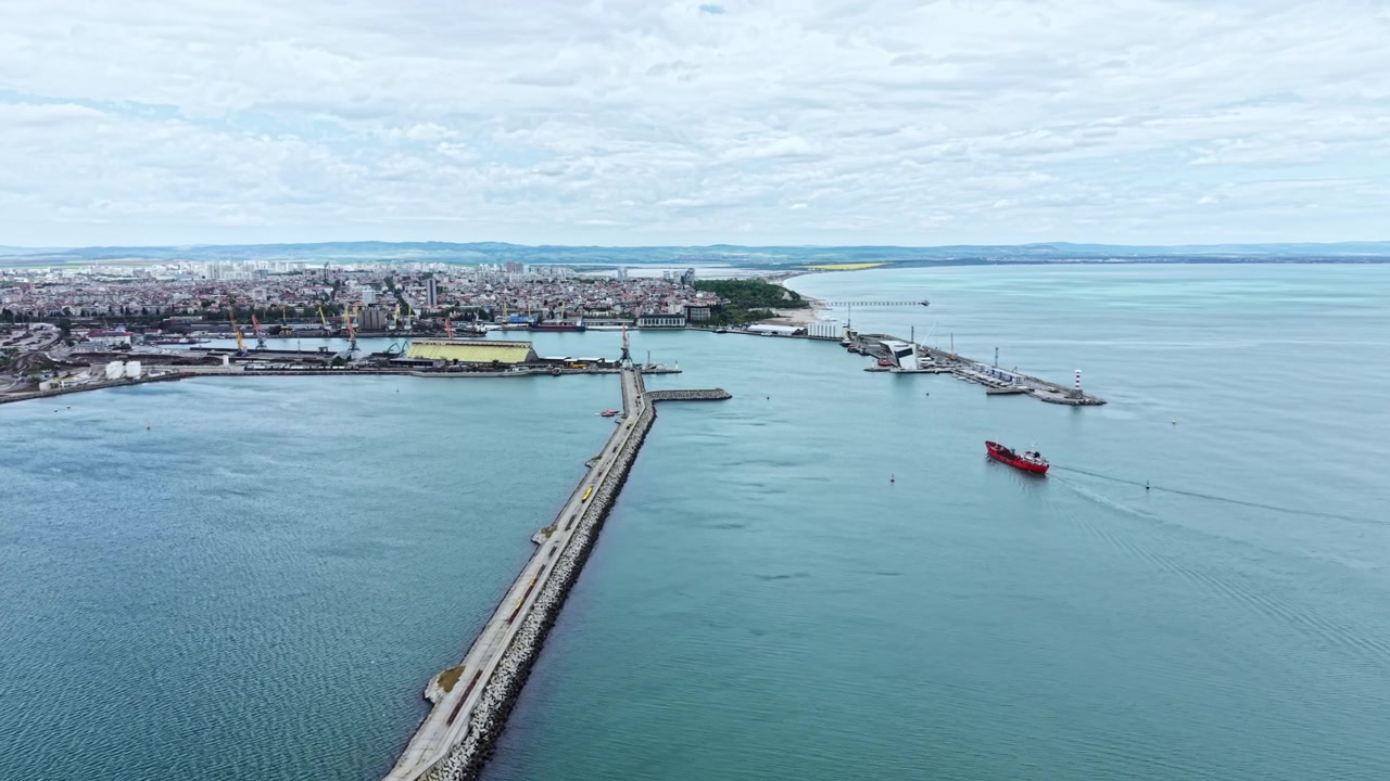 Vista di un porto dall'alto mostra una barca che naviga vicino alla costa con edifici e strutture industriali sullo sfondo durante tempo nuvoloso.