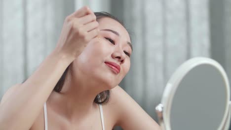 아름다운 젊은 아시아 여성이 집에서 메이크업하는 동안 세럼을 적용하고 미소를 짓는 거울을 봅니다.