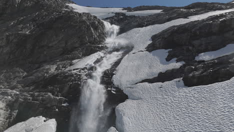 slowmotion drone shot flying around a powerfull waterfall near langvatnet lake in norway close to the strynefjellsveg breaking through ice and snow on a sunny day surrounded by shiny rocks log