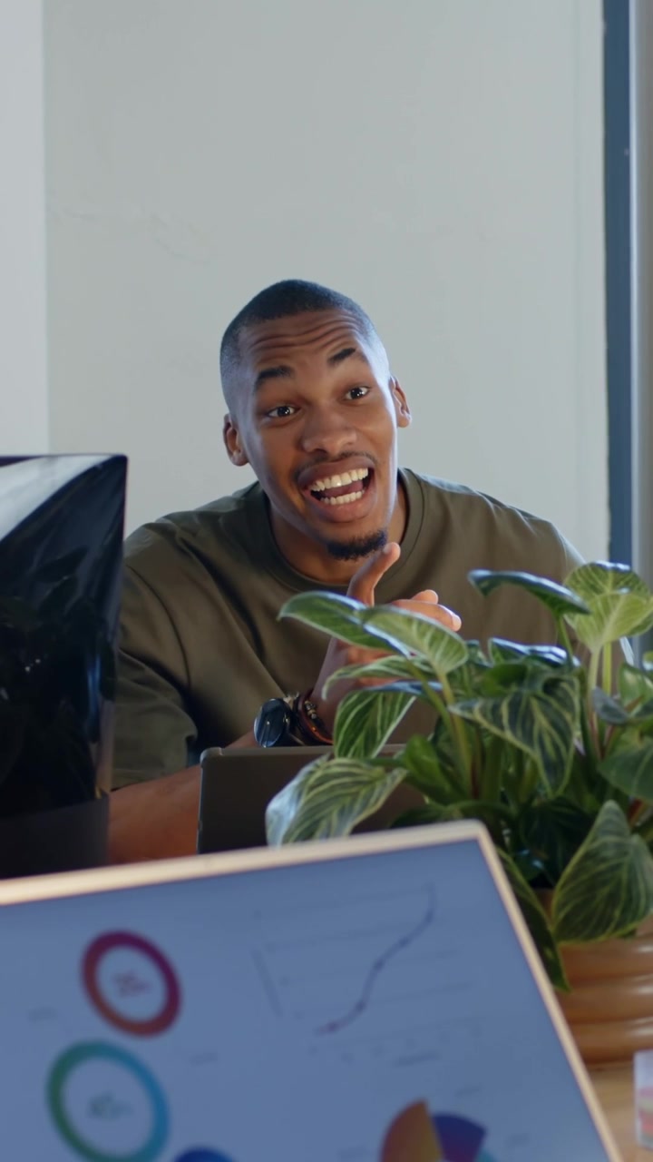 Video verticale: Uomo sorridente con maglietta color oliva che reagisce a un commento di un collega, appoggiato a un tavolo con un laptop.