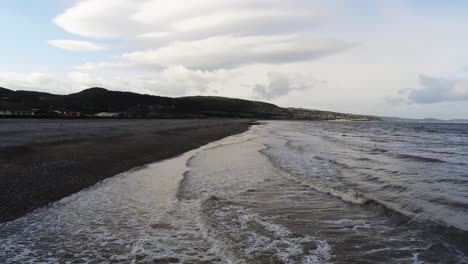 Aerial-view-following-shoreline-above-Pensarn-stone-beach-ocean-waves-under-Welsh-mountains