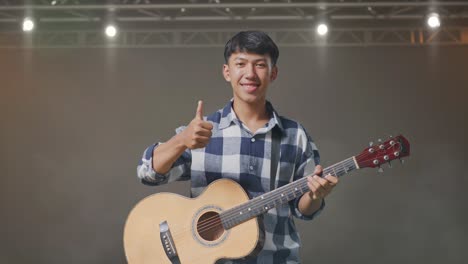 adolescente asiático con guitarra mostrando el pulgar hacia arriba y sonriendo en el escenario