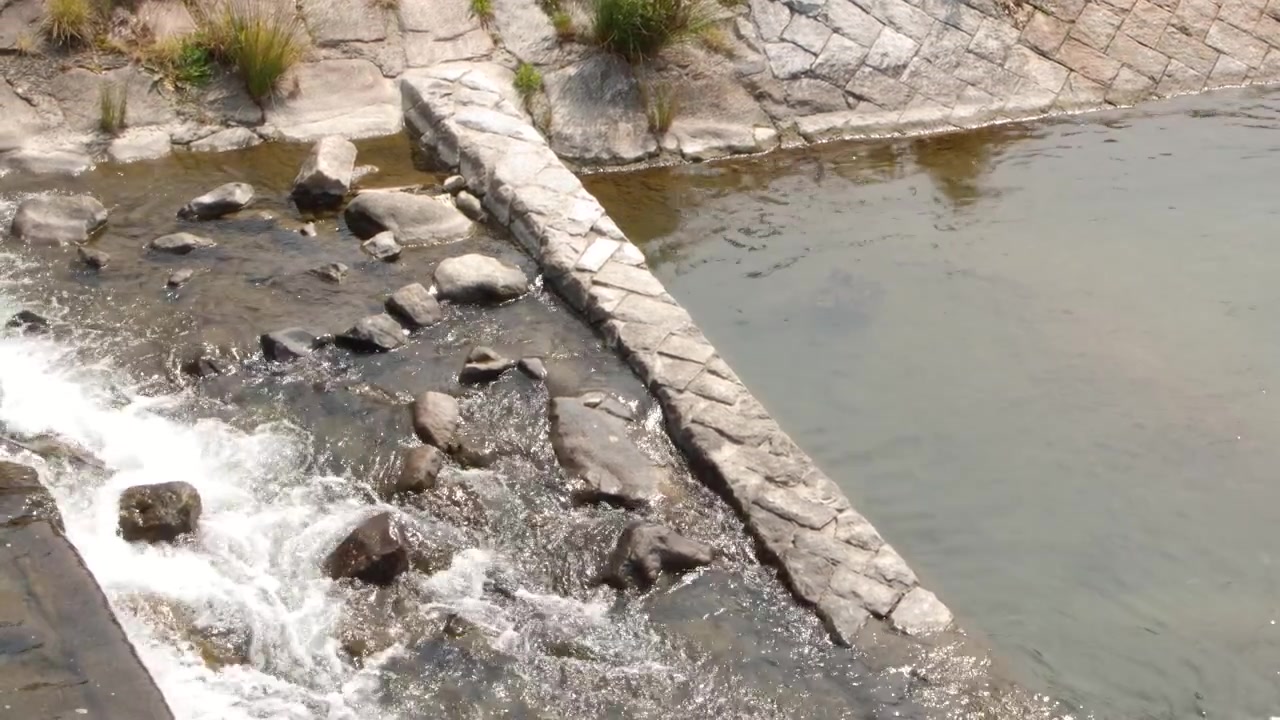 Toma aérea de agua de río precipitándose a través de una alcantarilla de hormigón y un terraplén de piedra.