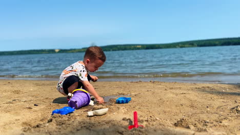 serious baby boy sits squatted on the beach. toddler plays with toy in the sand near the river.