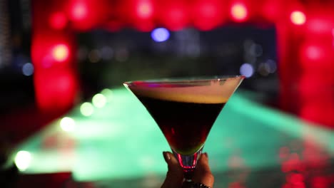 a hand swirls an espresso martini in a martini glass at a neon-lit bar, with colorful bokeh lights and shallow depth of field