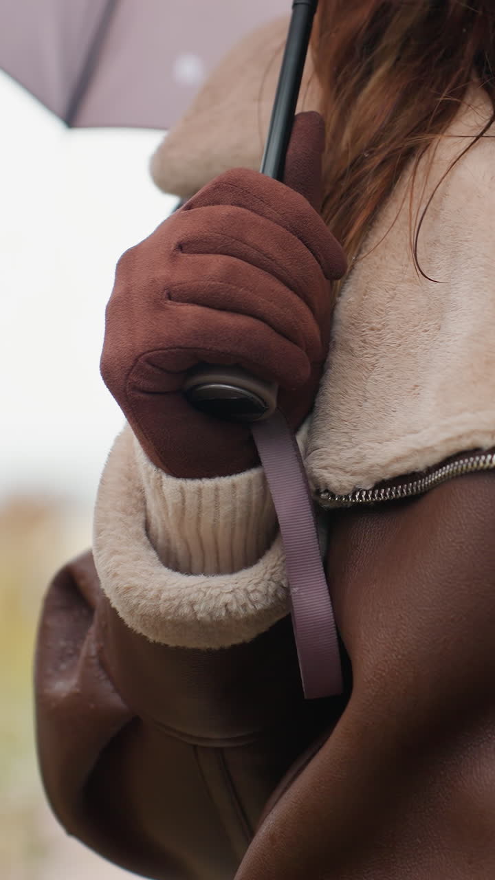 Lady with face partially visible, wearing brown jacket and brown gloves, holding umbrella, with soft focus background of nature and urban setting, creating a cozy, seasonal vibe