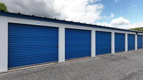 Aerial-view-of-a-row-of-blue-storage-units-with-roll-up-doors-in-a-secure-facility,-set-against-a-bright,-clear-sky