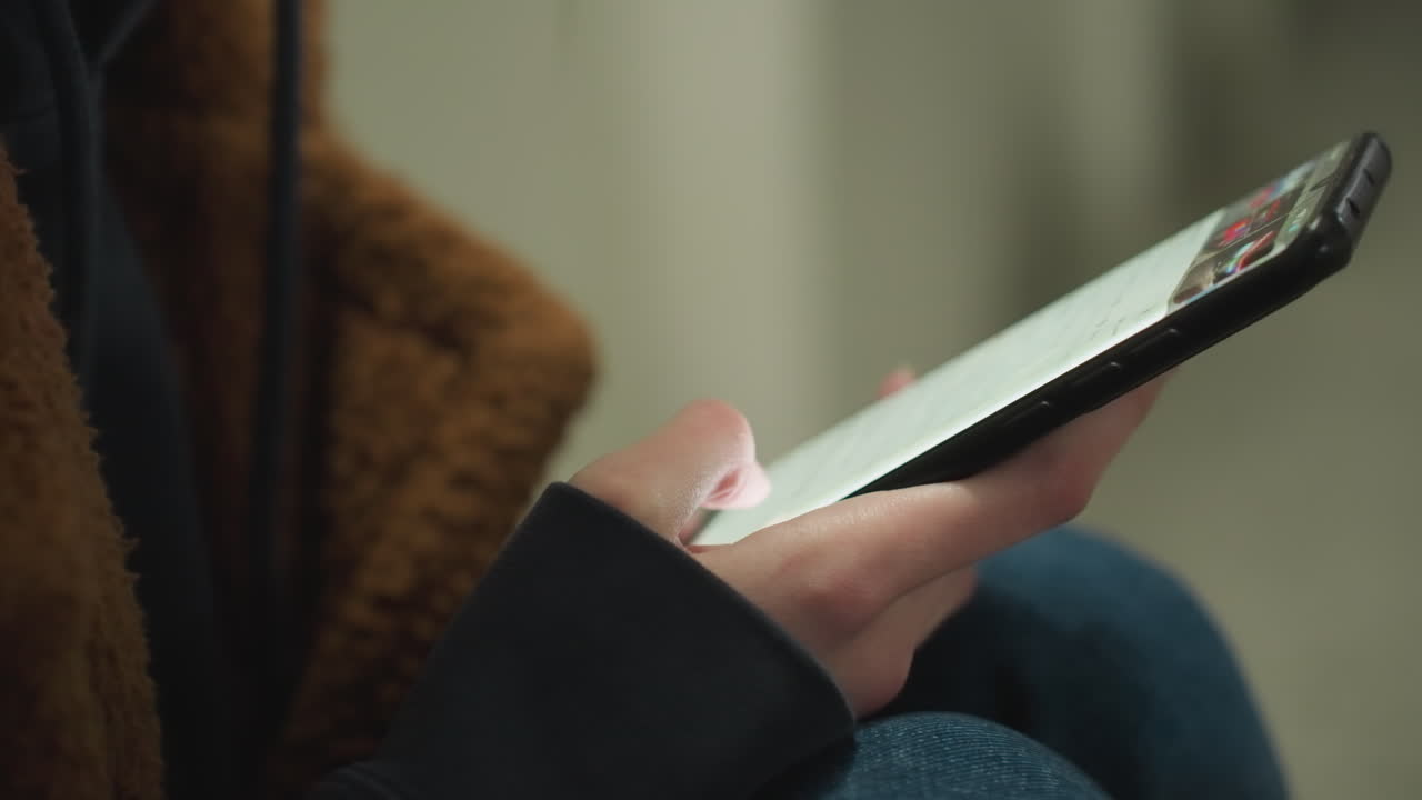 primer plano de una mano con un abrigo marrón, sosteniendo y desplazándose por la pantalla de un teléfono inteligente. la toma captura el enfoque en el dispositivo, transmitiendo una sensación de soledad, compromiso y contemplación