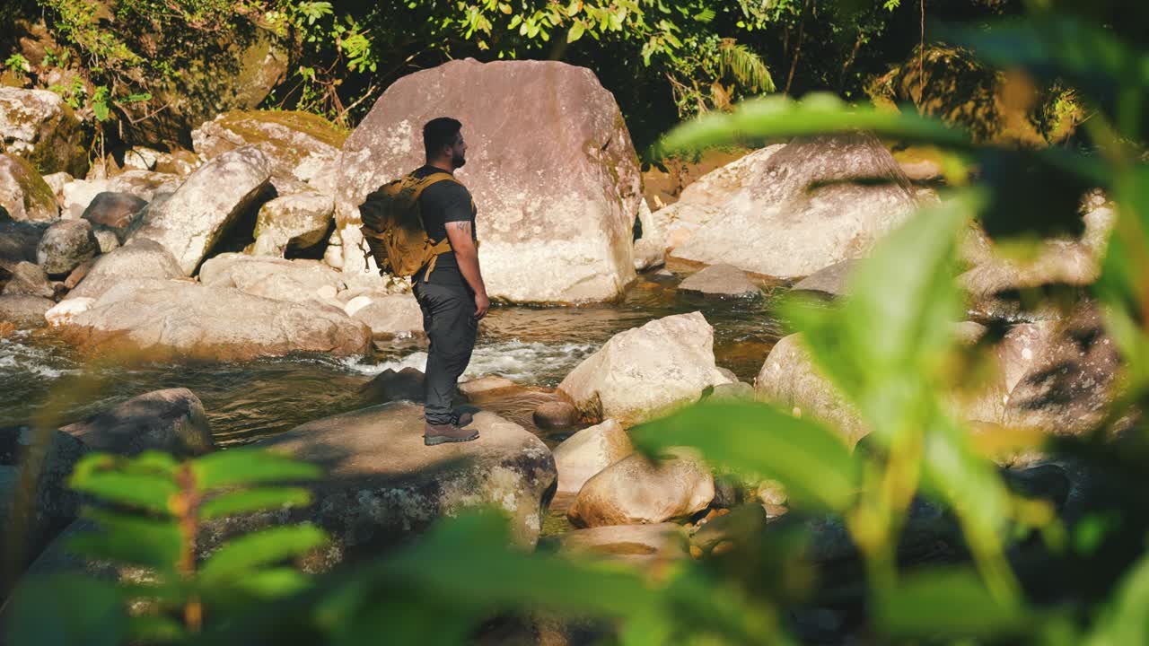 산악 강을 따라 오르는 남성 하이커는 바위와 나무로 둘러싸여 정글에서 하이크하는 동안 주위를 둘러보고 있습니다.