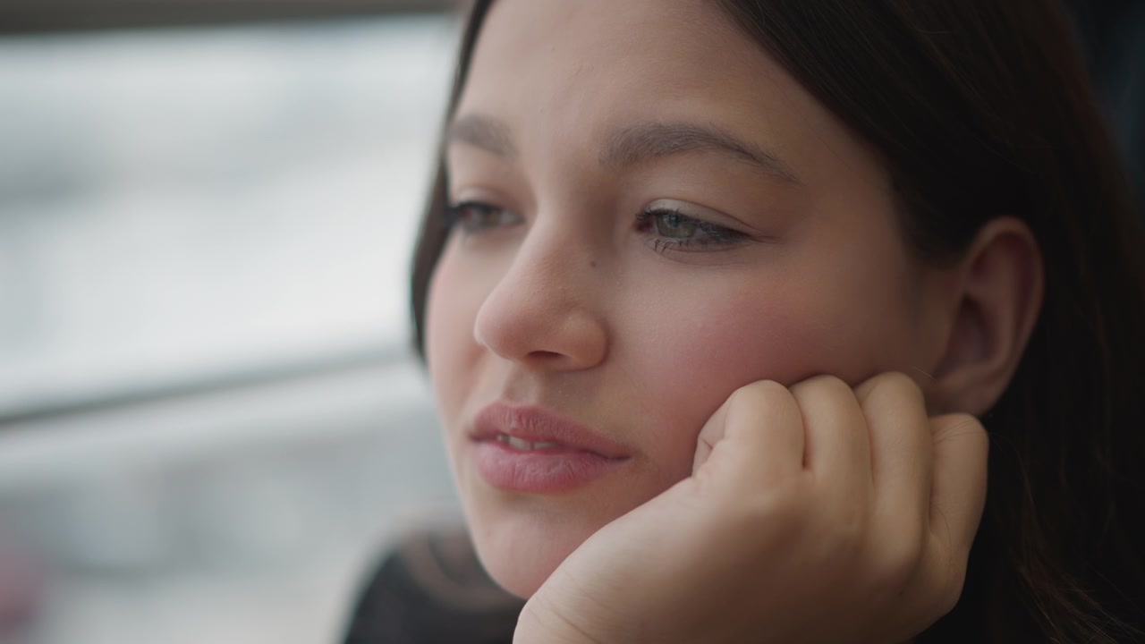Close portrait of teenage girl turning from contemplative gaze to mild amusement while resting chin on hand, touching cheek, then smoothing hair near ear in soft light and calm interior