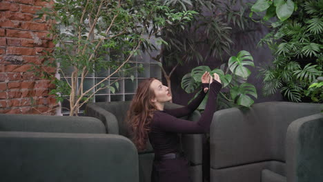 Lady raises phone for selfie while seated in modern booth adorned with lush indoor plants and brick walls, capturing moment in cozy green lounge with natural design elements and soft cushions