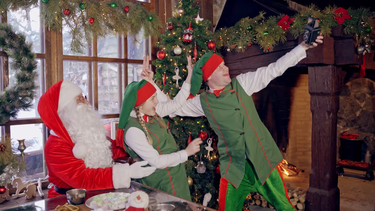 Funny elves with Santa Claus. Fairy characters doing selfie on the Christmas background indoors. Joyful atmosphere at New Year time.
