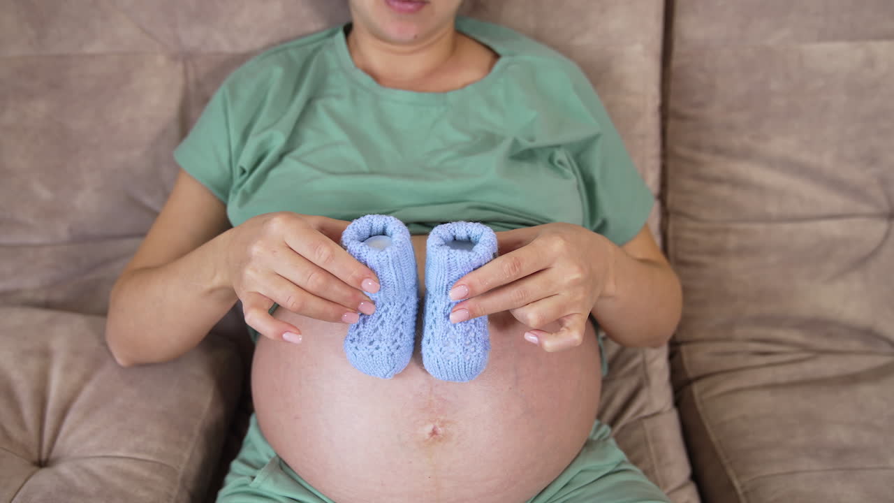 Future mum playing by little socks on her big belly. Pregnant woman with blue knitted socks on a her bare stomach. Top view.