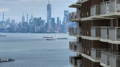 Aerial-revealing-shot-of-apartment-block-with-balcony-and-beautiful-american-Skyline-with-skyscraper-buildings-in-background---Ships-cruising-on-Upper-bay---New-York-City,America