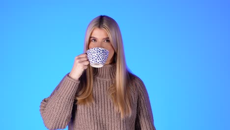 Porträt-Einer-Hübschen-Blonden-Frau,-Die-Aus-Einer-Kaffeetasse-Vor-Blauem-Hintergrund-Trinkt,-Studioaufnahme