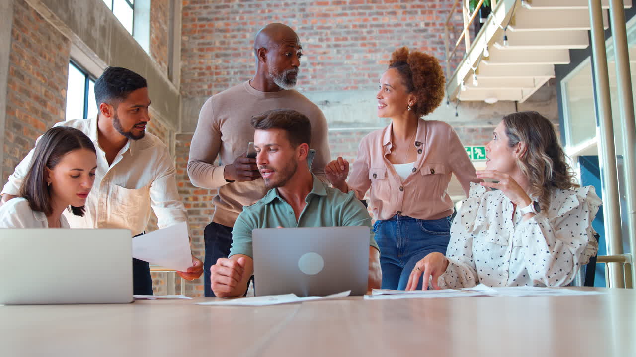 Multi-Cultural Business Team Meeting Around Laptop Discussing Documents In Busy Office