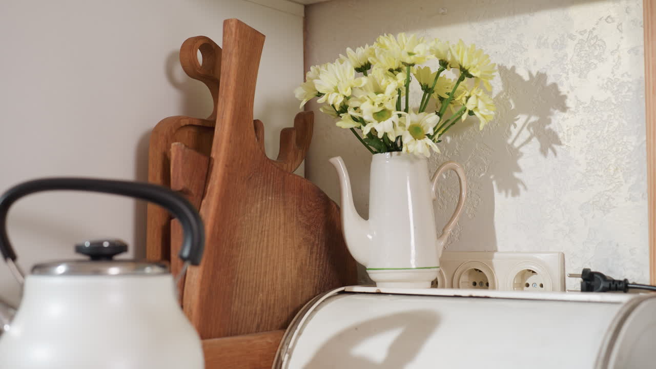 White electric kettle on kitchen counter next to wooden chopping boards, ceramic jug with flowers, hand blender, and storage canister, all arranged in cozy, naturally lit modern rustic kitchen