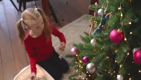 vista superior de la niña rubia arrodillada en el suelo recogiendo la decoración navideña de una caja y colgándola en el árbol de navidad 1