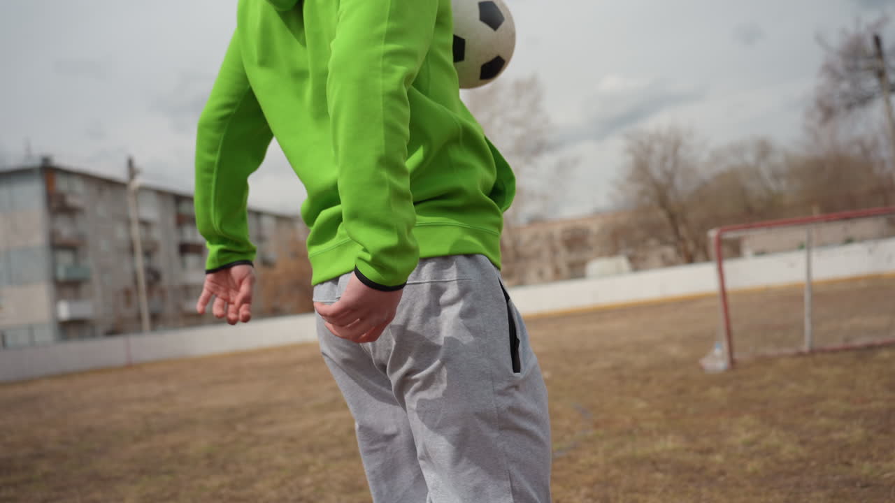 후면 시점에서 바라본 백인 운동선수가 탁 트인 도시 필드에서 축구공을 차올리는 모습이에요. 초록색 후드티와 회색 바지를 입고 있으며, 흐린 수평선과 아파트 단지가 보입니다. 장난기 넘치는 로프티드 패스와 혼자서 훈련하는 에너지가 느껴집니다.