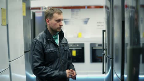 Young-man-is-choosing-a-refrigerator-in-a-store.-He-is-opening-the-doors,-looking-inside