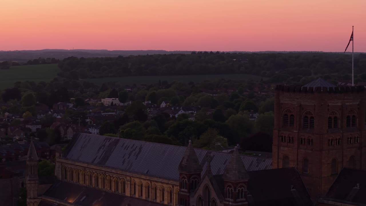 Aerial drone pull back over St Albans reveals the majestic cathedral glowing in golden hour light, with a soft pink horizon and the peaceful English landscape