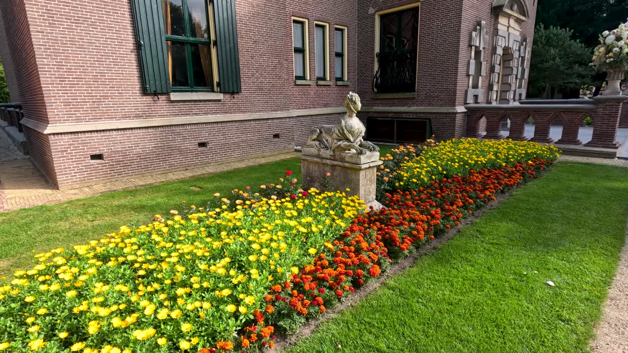 Camera glides alongside vibrant flower beds and a historic brick building with statues, capturing lush gardens in bright natural daylight at a botanical site