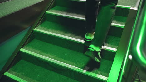 close-up of man's foot in black and white sneaker stepping up stairs under green lighting. metal railings and carpeted steps create a sleek, modern look with a futuristic ambiance