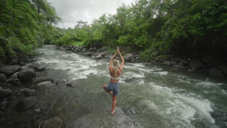 cuerpo tonificado femenino practicando la pose del árbol vrikshasana en una roca al lado del río, bali