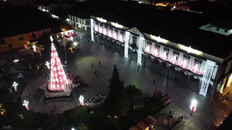 Toma-En-ángulo-Alto-Sobre-El-Palacio-De-Gobierno-En-El-Pequeño-Pueblo-Navideño-En-El-Centro-De-México-Con-Gente-Caminando-Decorada-Con-Hermosas-Decoraciones-Navideñas-En-La-Noche