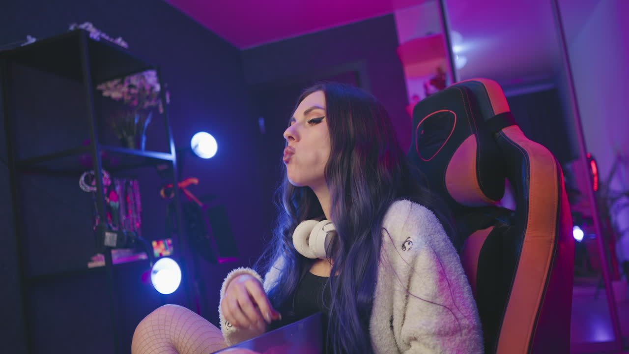 Fair skinned lady with long dark hair enjoying popcorn while seated in gaming chair, headphones resting around neck, surrounded by pink and purple studio lights with soft ambient glow