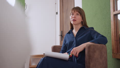 emotional coach seated comfortably on chair, talking with visitor in cozy therapy lounge, holding pen and notepad while engaging in thoughtful supportive conversation in calm interior setting