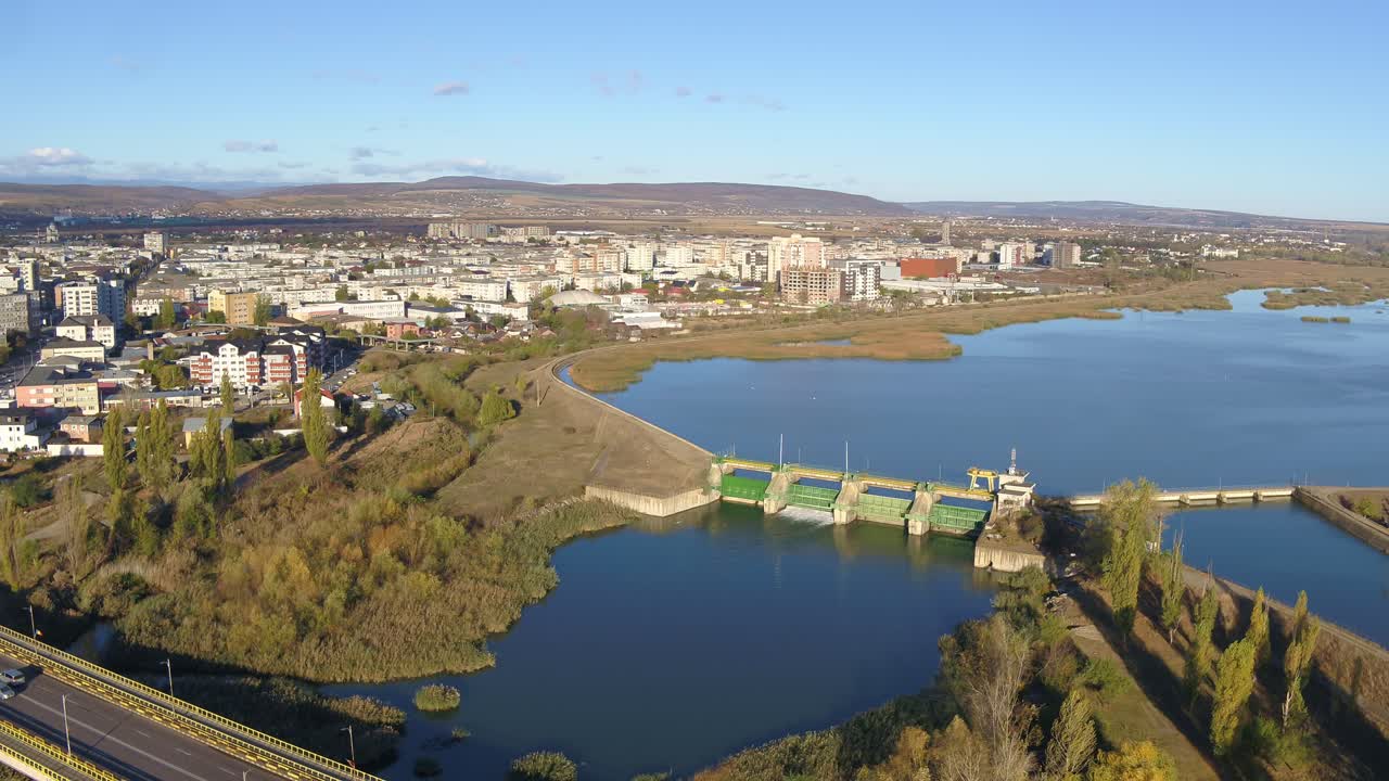 A slow-panning aerial shot shows a hydroelectric dam in Bacau with green sluice gates on a river. The reservoir sits beside a city on a beautiful sunny autumn day in Romania