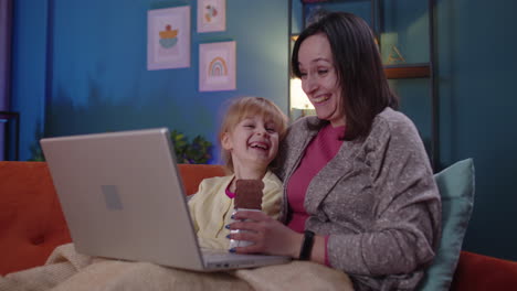 madre y niño pequeño hija niño estudio en la computadora portátil, viendo películas divertidas, relajándose, acostado en el sofá