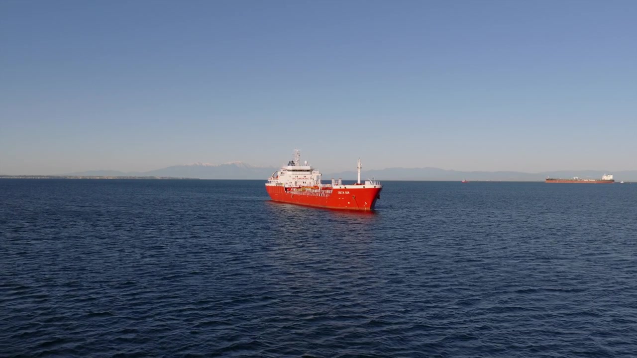 Un buque de carga naranja navega por las aguas del golfo cerca de Tesalónica, con otro barco visible a lo lejos.