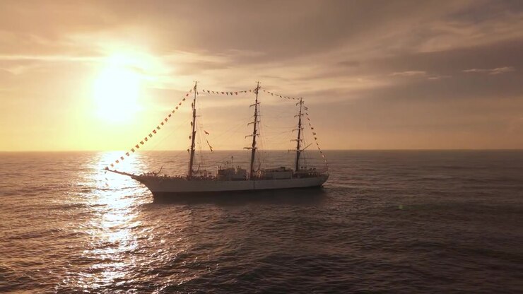 Aerial lateral tracking shot of a three-masted tall ship sailing on the open ocean during a golden sunset, featuring heavy backlighting and sun reflections on the water