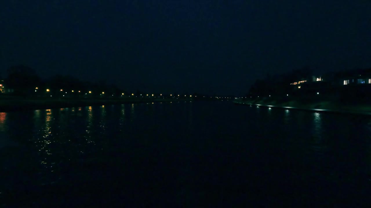 La camera mostra un fiume di notte. Le luci degli edifici e delle lampade si riflettono sull'acqua. L'area circostante è buia poiché il cielo notturno è sereno