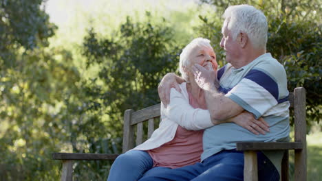 happy senior caucasian couple sitting on bench embracing in sunny garden, slow motion