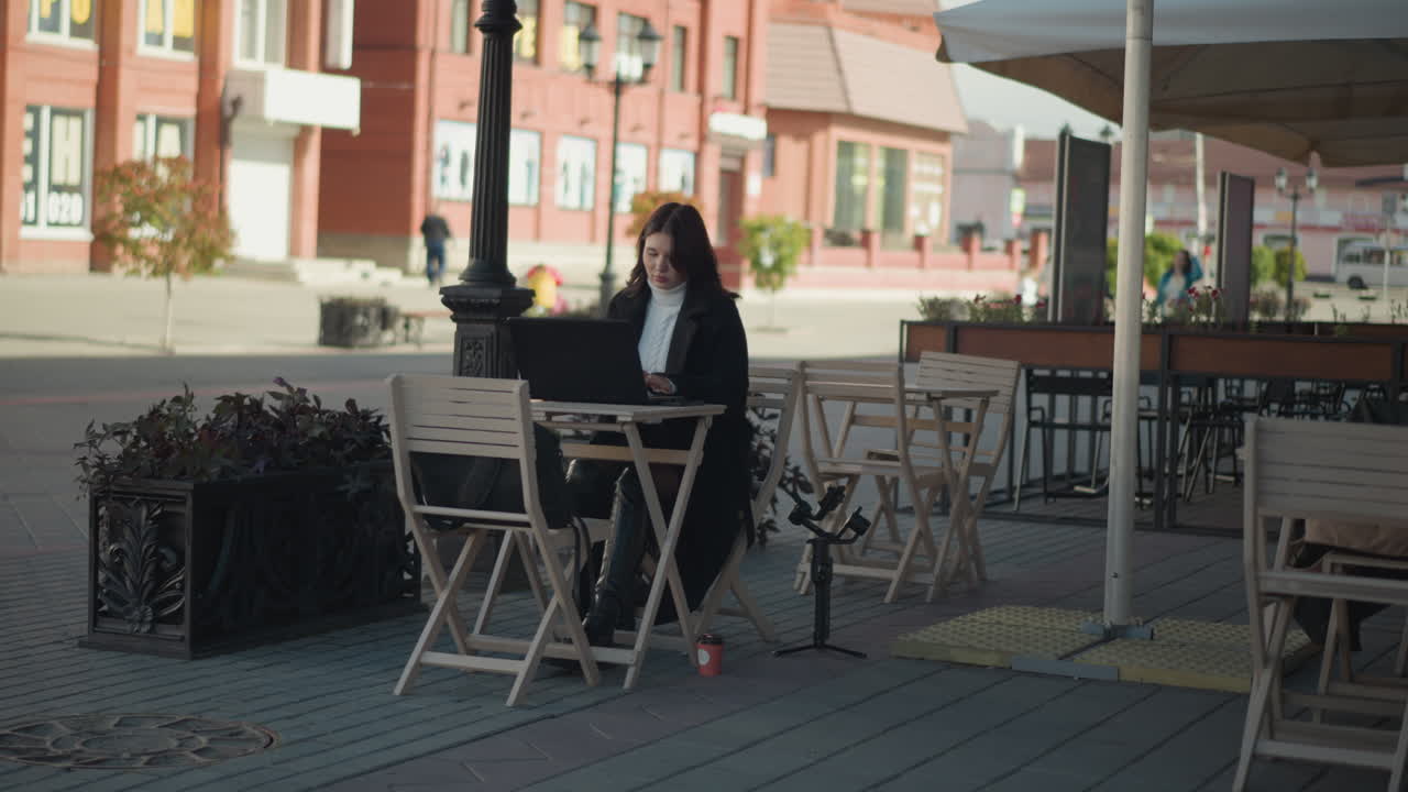 mujer de cabello oscuro trabajando en una computadora portátil en una mesa al aire libre en un ambiente acogedor de cafetería, rodeada de plantas y sillas con un fondo borroso que muestra la calle de la ciudad, el autobús pasando en la lejanía