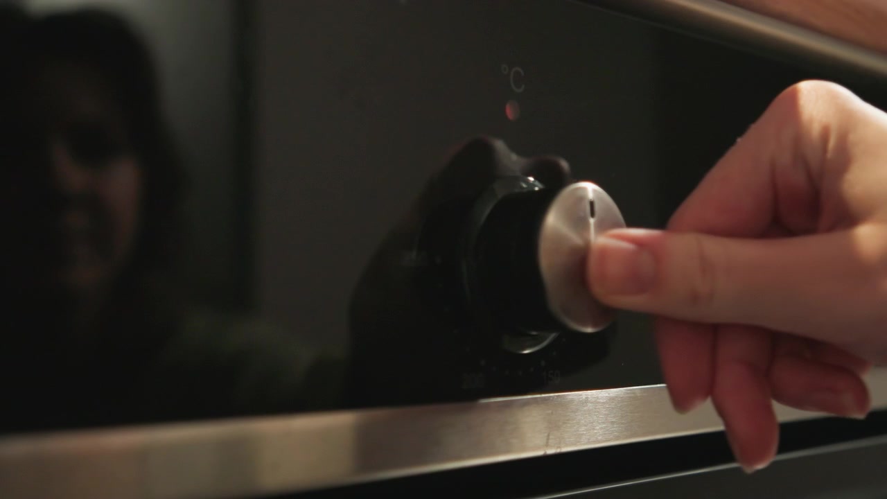 Una vista ravvicinata dal basso mostra una manopola rotante metallica che viene girata su un pannello di vetro scuro di un elettrodomestico, con contatto della punta del dito, rotazione visibile, display illuminato, finitura in acciaio inossidabile spazzolato e dettagli sottili.
