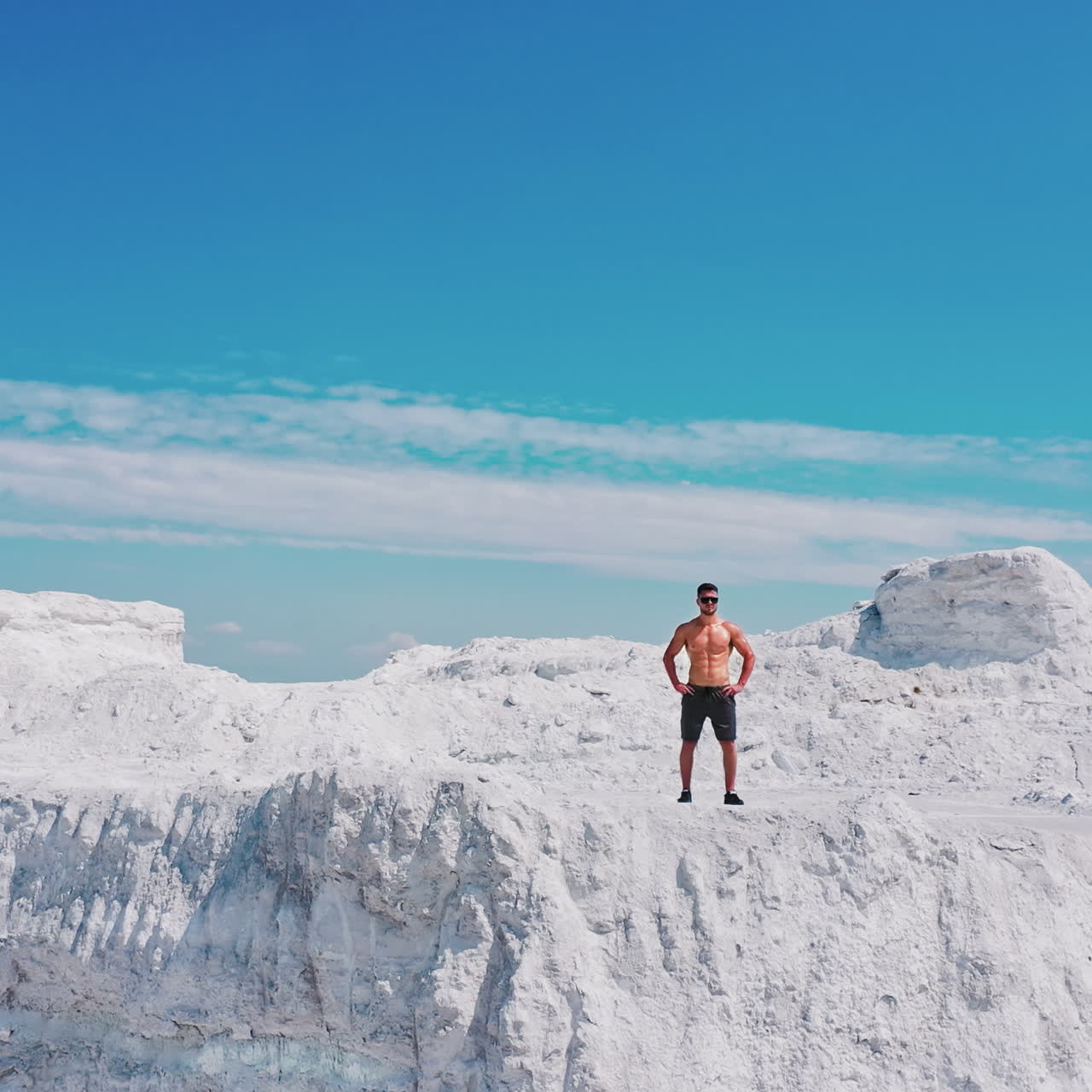 Sporty man with muscular body on the top of white hill under blue sky. Handsome sportsman on beautiful summer landscape in the mountains. Motion down.