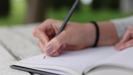 female-hands-writing-in-journal,-sitting-on-white-table-in-the-green-garden,-handheld-closeup