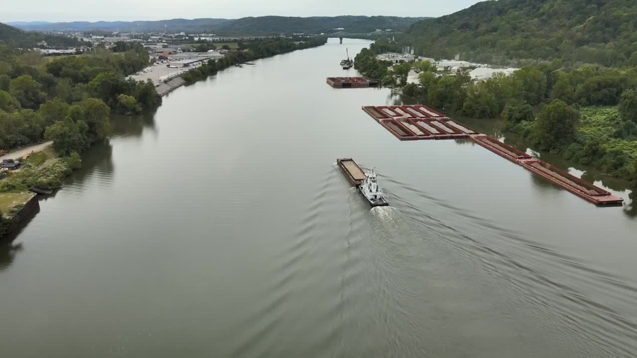 Aerial drone footage of towboat guiding cargo barge along calm Kanawha River surrounded by forested hills, industrial docks, and quiet roads in scenic West Virginia, United States