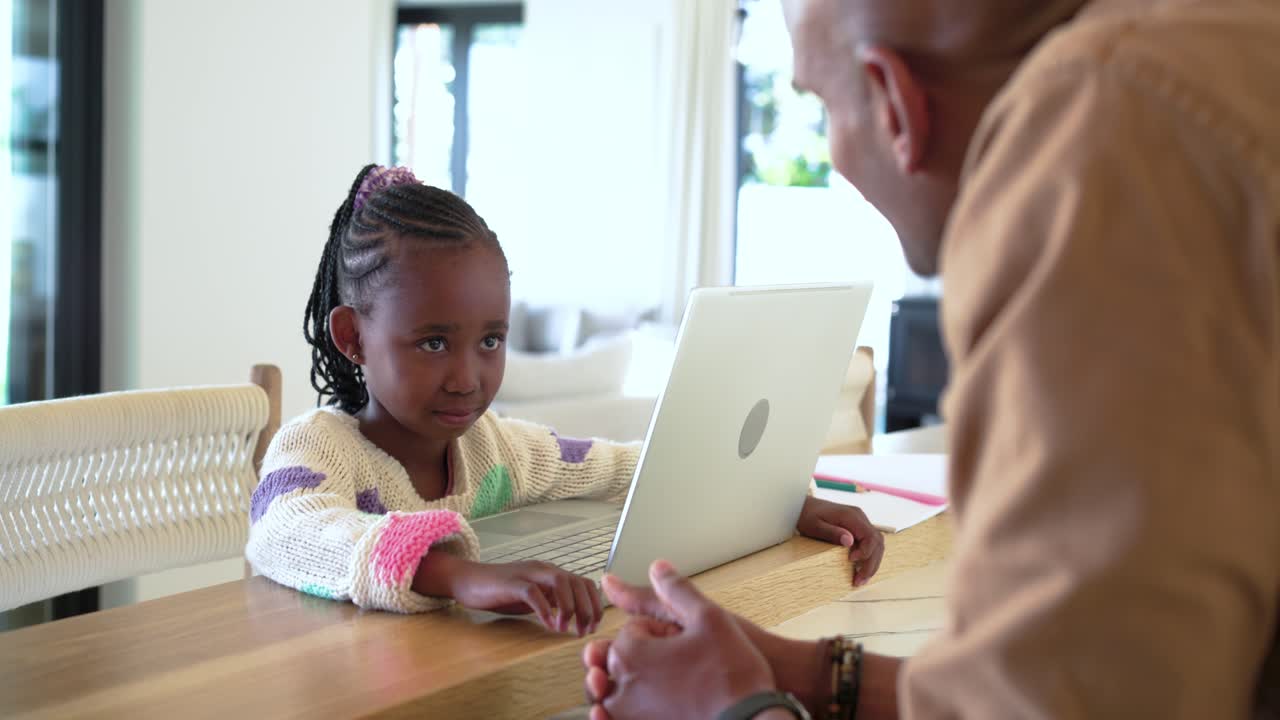 Ragazza afroamericana seduta al bancone della cucina con un laptop color argento, un uomo si sporge per guidarla