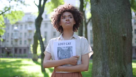 upset african-american woman looking around, holding missing cat poster in hands